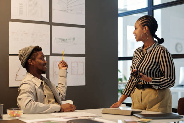 Young confident architect pointing at blackboard with sketches of new buildings and looking at female colleague during presentation
