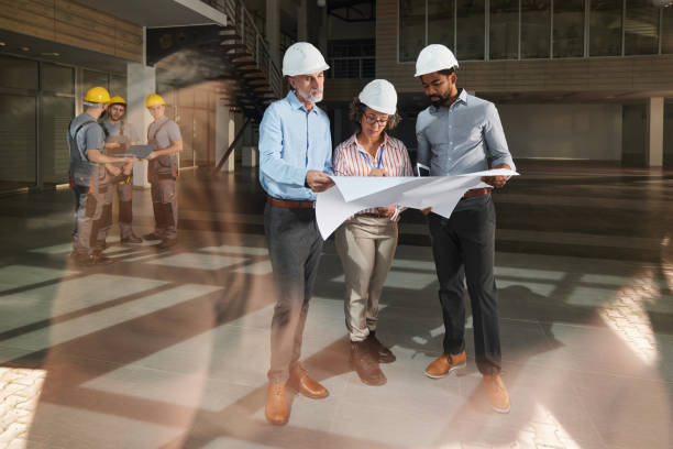 Team of diverse real estate developers cooperating while working on blueprints at construction building. Their manual workers are in the background. The view is through glass.