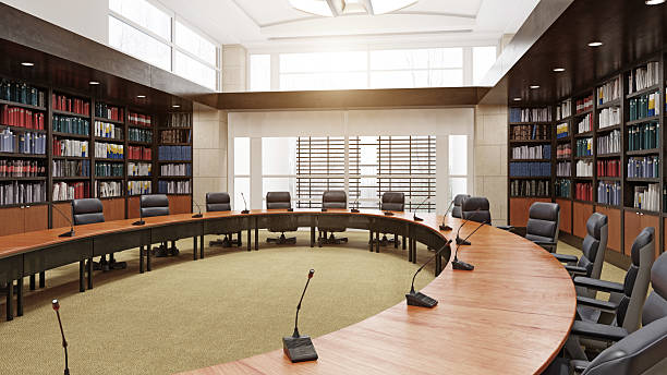 A 3D rendering of a modern, empty conference room with a large curved wooden table, black office chairs, and microphones. The room features floor-to-ceiling bookshelvesâ¦